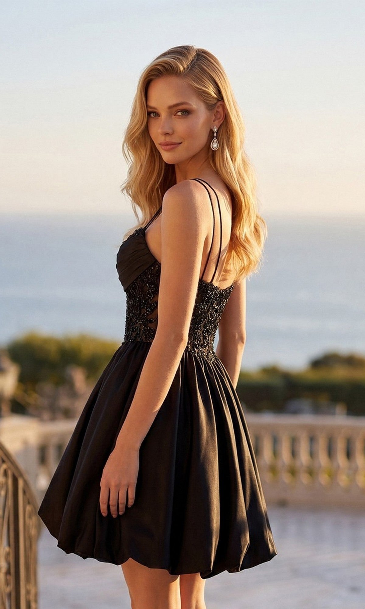 Wearing the Juliet JT7186A Short Homecoming Dress, a woman stands outdoors with the ocean and greenery behind her, glancing softly over her shoulder. The sleeveless black dress features a beaded bodice that catches the light, perfectly complementing its satin a-line skirt.